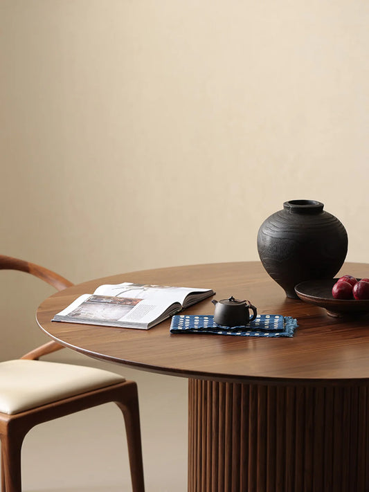 Angled view of a solid walnut round pedestal dining table with fluted base, styled with a black vase, fruit bowl, tea set, and open book.