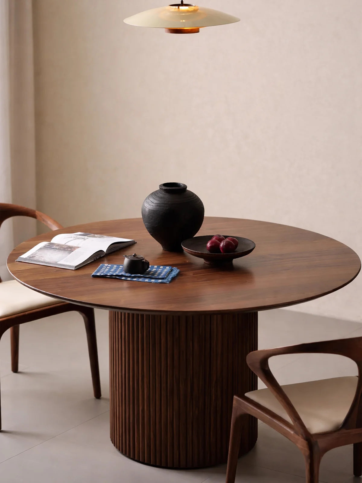 Angled front view of a solid walnut round pedestal dining table with a broad top, black vase, tea set, and fruit bowl.