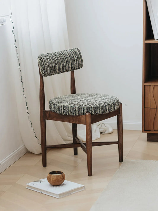 Eron upholstered dining chair in solid walnut, three-quarter side view in a bright room with curtains and walnut shelf