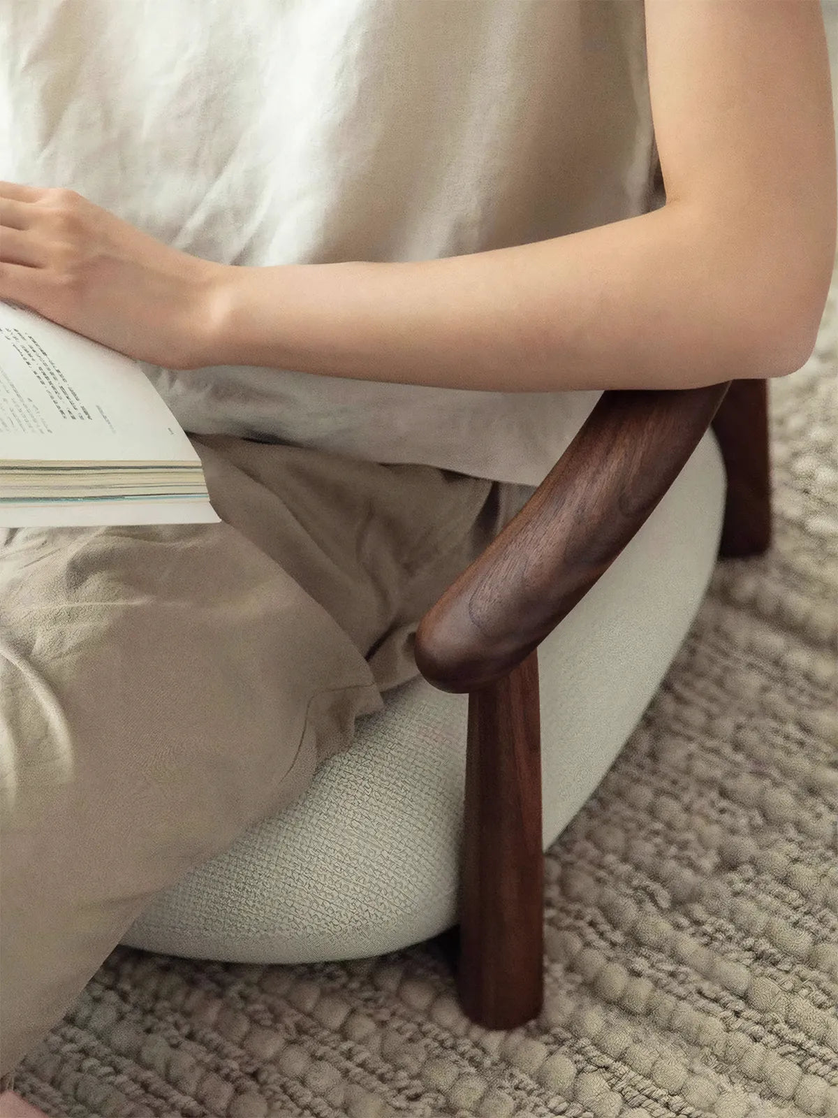 Close-up of a person reading comfortably in The Haven Wide-Seat Floor Chair, resting their arm on the smooth, hand-finished curved walnut armrest.