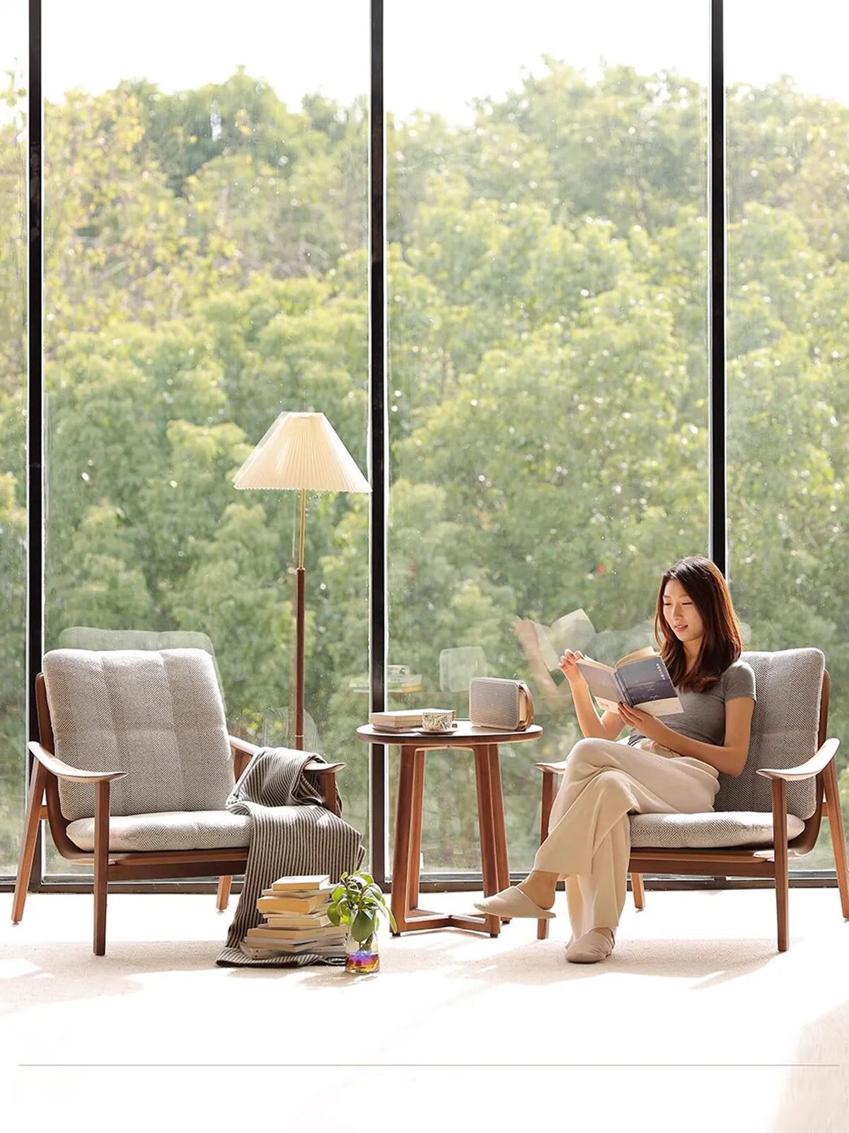 A woman reading a book in The Haven Reading Chair by Walnutry, arranged as a pair with a solid North American Black Walnut side table by a large sunlit window.