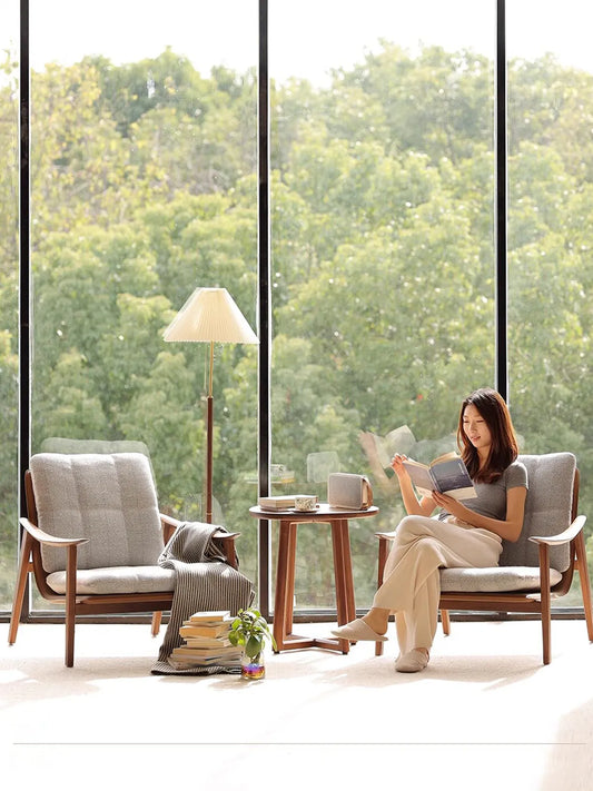 A woman reading a book in The Haven Reading Chair by Walnutry, arranged as a pair with a solid North American Black Walnut side table by a large sunlit window.
