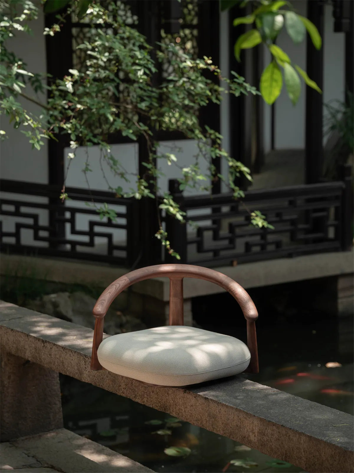 The Haven Wide-Seat Floor Chair crafted from rich dark walnut, resting elegantly on a stone ledge in a tranquil outdoor Zen garden.