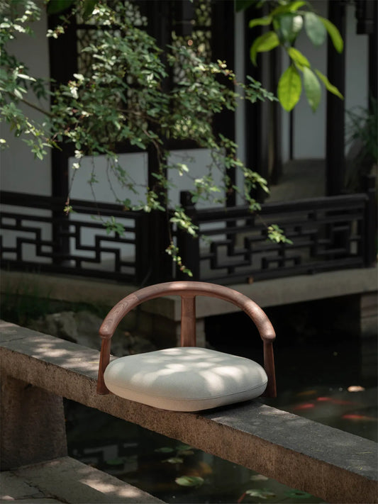 The Haven Wide-Seat Floor Chair crafted from rich dark walnut, resting elegantly on a stone ledge in a tranquil outdoor Zen garden.