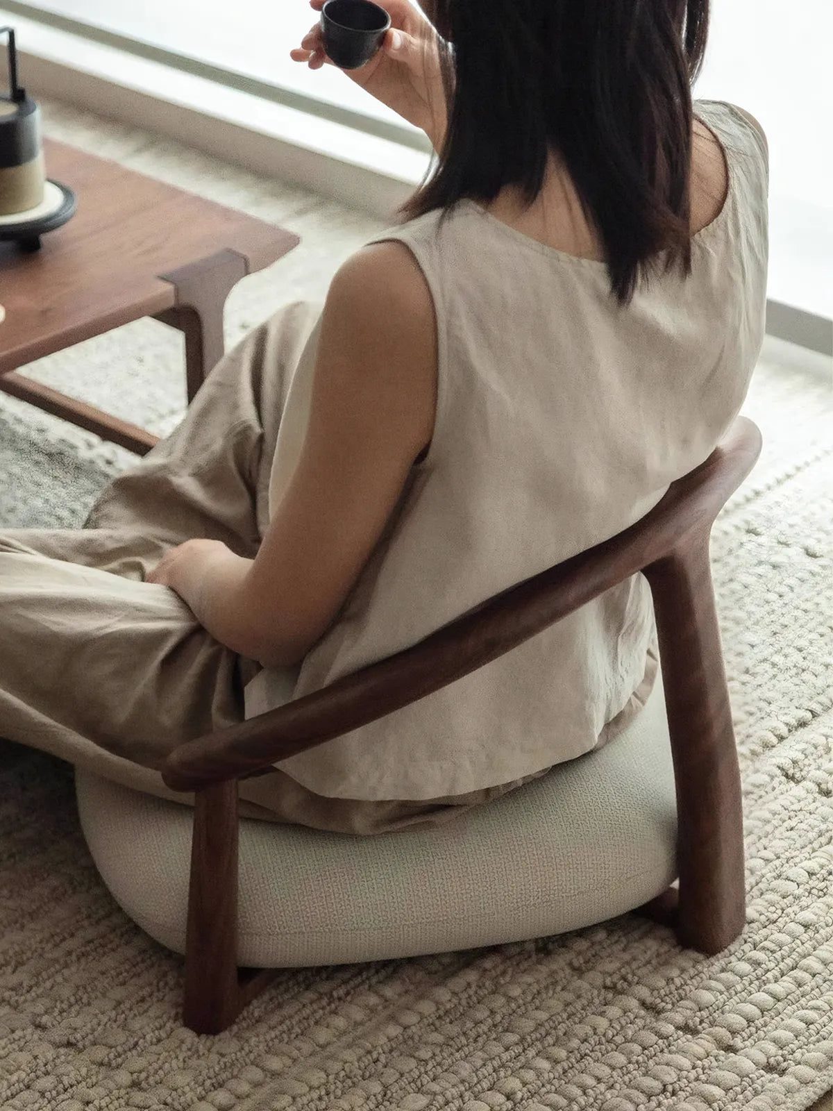 A woman enjoying tea while seated in The Haven Wide-Seat Floor Chair, demonstrating the ergonomic lower back support provided by the curved wooden frame.