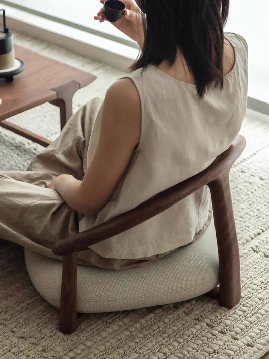 A woman enjoying tea while seated in The Haven Wide-Seat Floor Chair, demonstrating the ergonomic lower back support provided by the curved wooden frame.