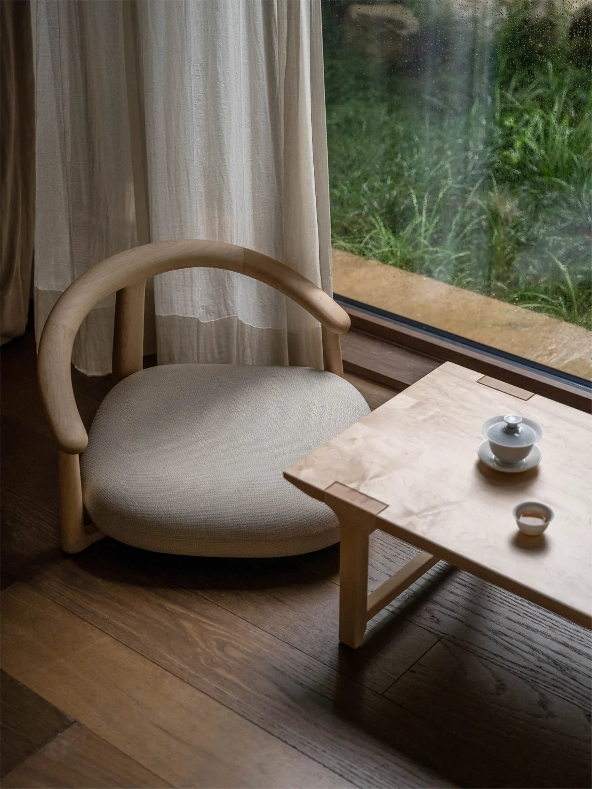 The Haven Wide-Seat Floor Chair styled in a serene, natural light wood finish next to a low tea table, overlooking a lush garden window.