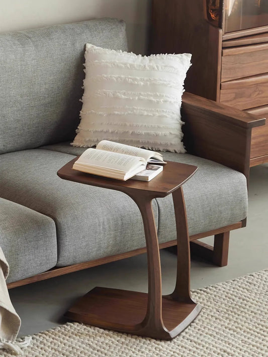 The Helios C-shaped side table conveniently pulled over a gray sofa, holding an open book in a cozy living room setting.