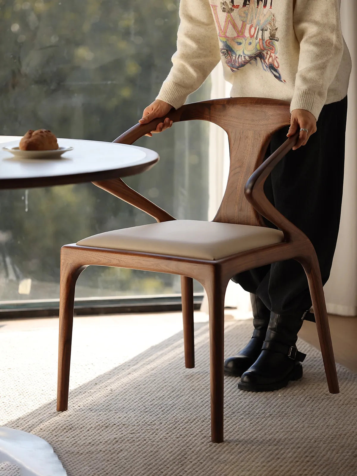 A person positioning the Helios walnut chair at a dining table, demonstrating the chair's scale and smooth finish.