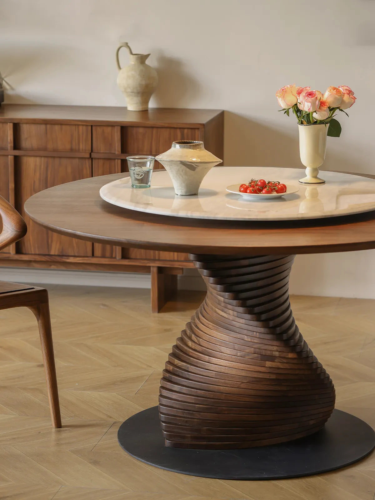 The striking spiral base of The Helios Round Dining Table highlighted by warm lighting, perfectly paired with a matching Walnutry solid wood sideboard in the background.
