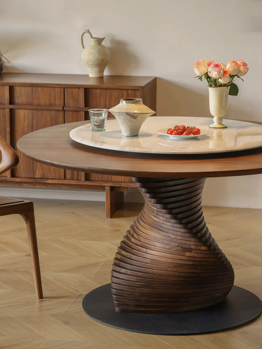 The striking spiral base of The Helios Round Dining Table highlighted by warm lighting, perfectly paired with a matching Walnutry solid wood sideboard in the background.