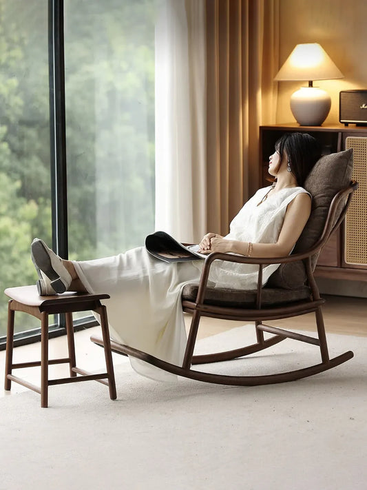 A woman relaxing comfortably in The Helios Rocking Chair by Walnutry, featuring a solid North American Black Walnut frame and a matching footstool by a large bright window.