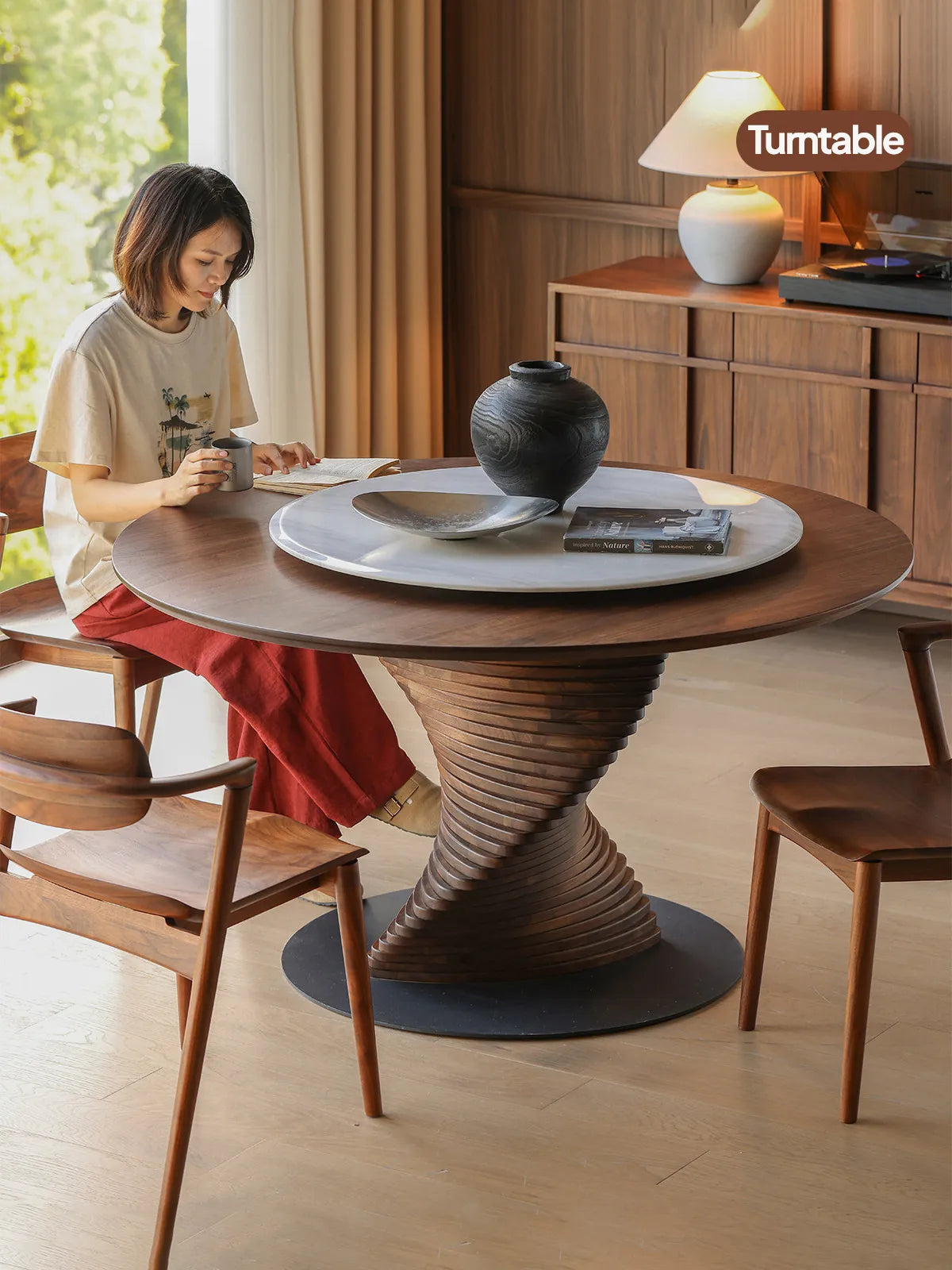 A woman relaxing and reading at The Helios Round Dining Table, demonstrating the scale of the table and the practical use of the integrated marble turntable.
