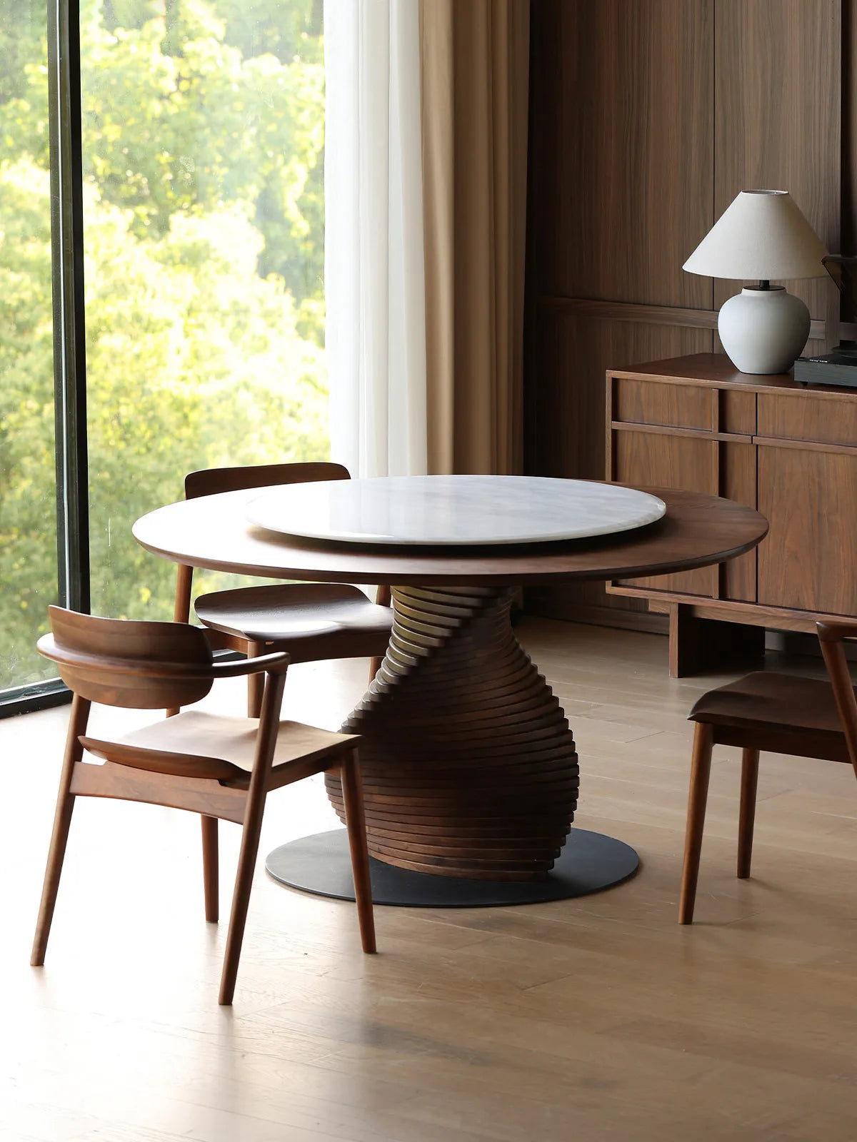 The Helios Round Dining Table and solid walnut dining chairs beautifully illuminated by bright natural light next to a large floor-to-ceiling window.