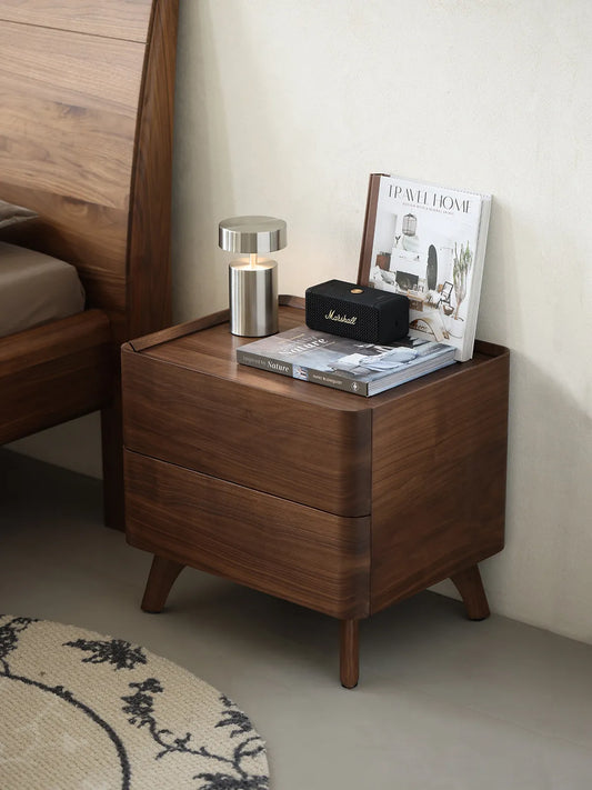 The Helios Solid Black Walnut Nightstand by Walnutry, styled next to a wooden bed with a modern lamp and speaker in a cozy bedroom.