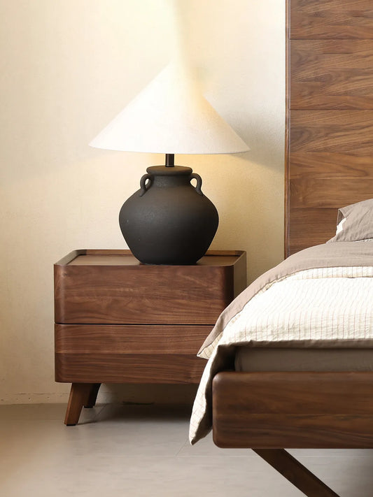 The Helios solid wood bedside table elegantly styled with a large decorative black lamp, adding sophisticated contrast to the natural walnut finish.