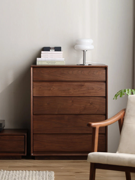 Front view of The Heritage solid walnut 5-drawer tallboy dresser, decorated with a modern white lamp and books in a bedroom setting.