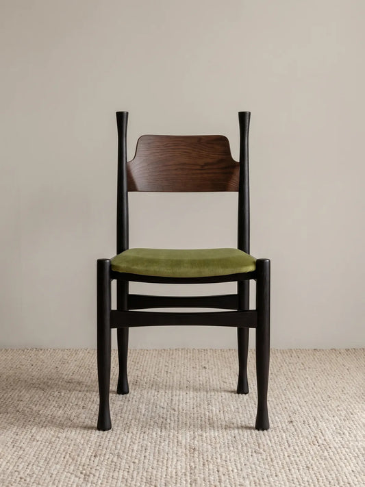 Korin dining chair front view showing black-finished frame, curved walnut backrest, and green velvet upholstered seat on a neutral rug