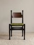 Korin dining chair front view showing black-finished frame, curved walnut backrest, and green velvet upholstered seat on a neutral rug