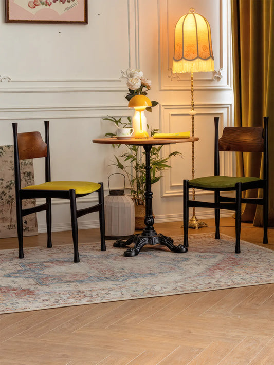 Two Korin dining chairs with green and yellow velvet seats flanking a small round table in a styled room with warm lighting