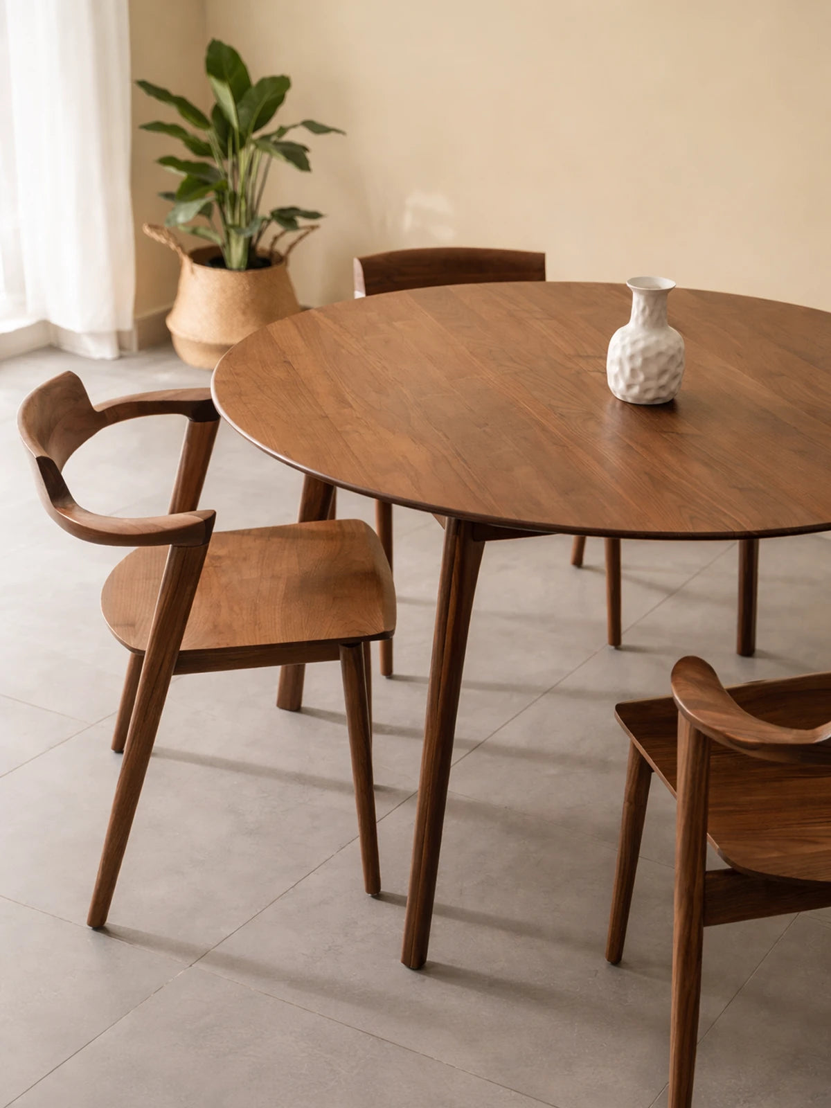 Round walnut dining table styled with wooden dining chairs, shown in a bright interior with a white vase on top and a potted plant in the background.