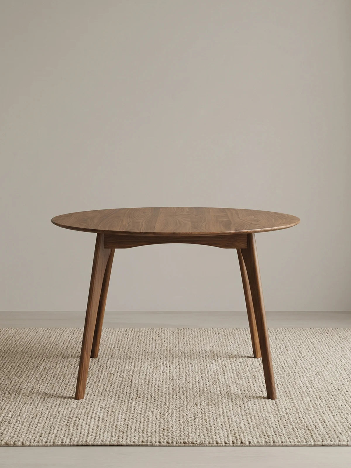 Studio view of a round walnut dining table with angled legs, shown on a textured neutral rug against a plain backdrop.