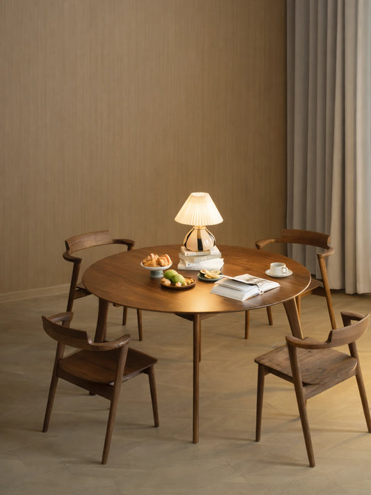 Round walnut dining table styled with four matching wooden chairs, a table lamp, fruit, pastries, and an open book in a warm dining room setting.