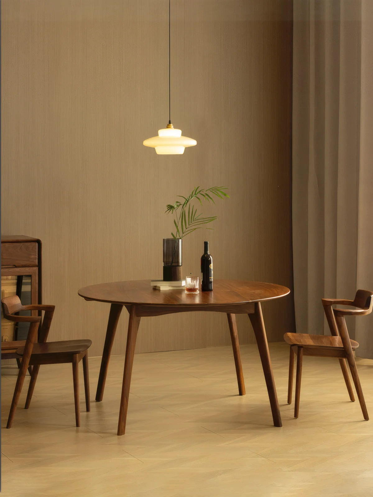 Round walnut dining table in a warm interior with a pendant light overhead, styled with a vase, wine bottle, and glass, and surrounded by matching wooden chairs.