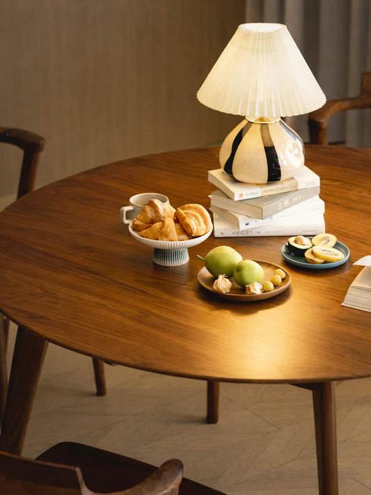 Close-up of the round walnut dining table styled with a table lamp, pastries, fruit, books, and a coffee cup.
