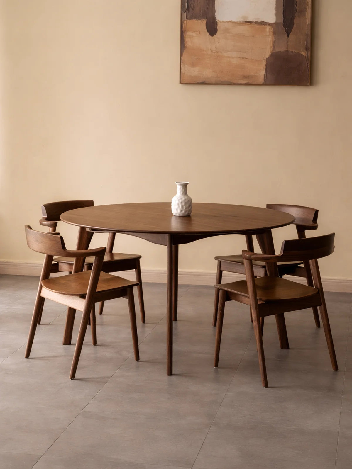 Front view of a round walnut dining table with four matching wooden chairs, styled with a small white vase in a minimalist dining space.
