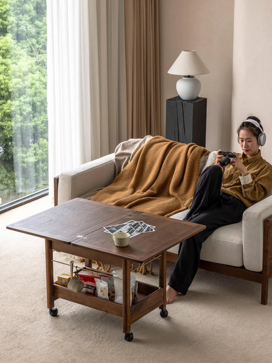A woman relaxing on a living room sofa, using the expanded Nomad Mobile Folding Side Table by Walnutry to conveniently hold a mug and documents.