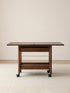 Front-facing view of the fully extended Nomad Mobile Folding Side Table, displaying its solid black walnut frame, smooth caster wheels, and lower storage rack.