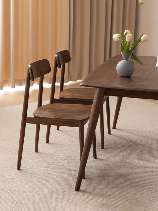 Two solid walnut dining chairs paired with a matching dining table in a calm interior, showing the chair’s clean proportions and warm wood tone.