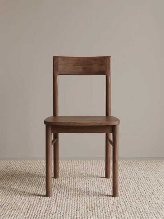 Riven solid walnut dining chair seen from the front, showing low curved backrest, contoured wood seat, and rounded stretcher bars