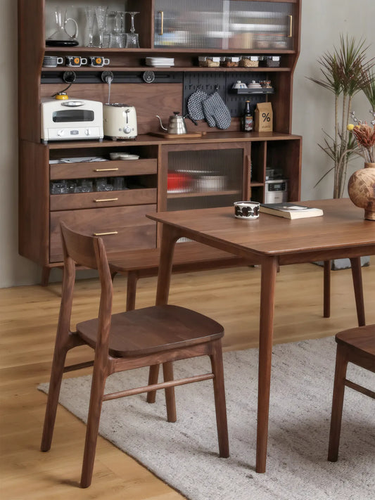 Side lifestyle view of the Riven walnut dining chair at a table with a walnut sideboard, mug, and book in the background
