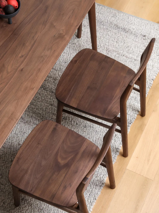 Overhead view of two Riven walnut dining chairs tucked under a walnut dining table, showing broad wood seat grain and open backrest