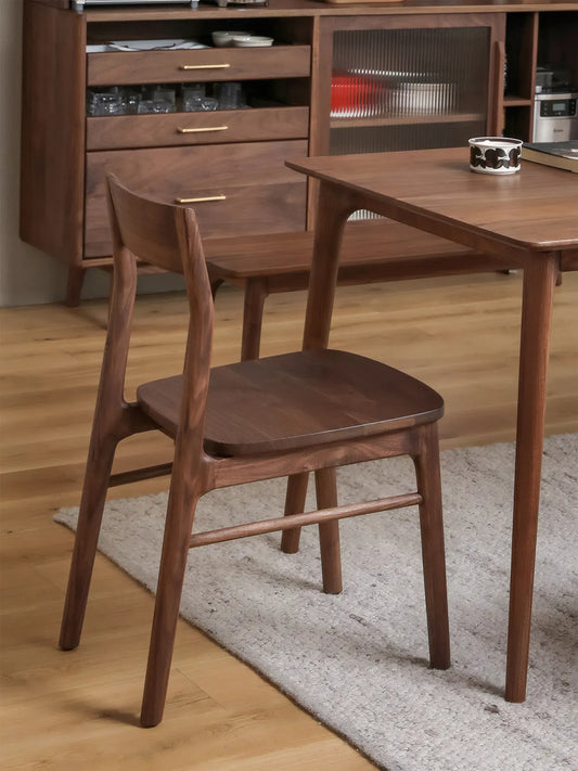Rear three-quarter view of the Riven walnut dining chair beside a dining table, showing curved backrest and tapered back legs