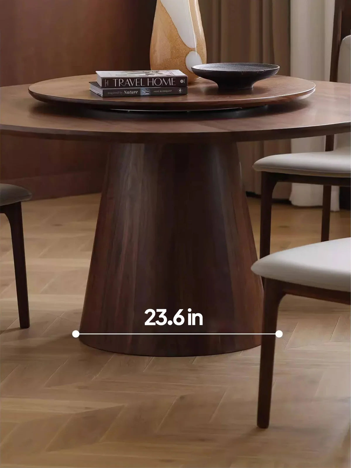Close-up of the tapered pedestal base on a dark walnut round dining table, showing a 23.6-inch base width, with a matching lazy Susan, books, a vase, and a black bowl on top.
