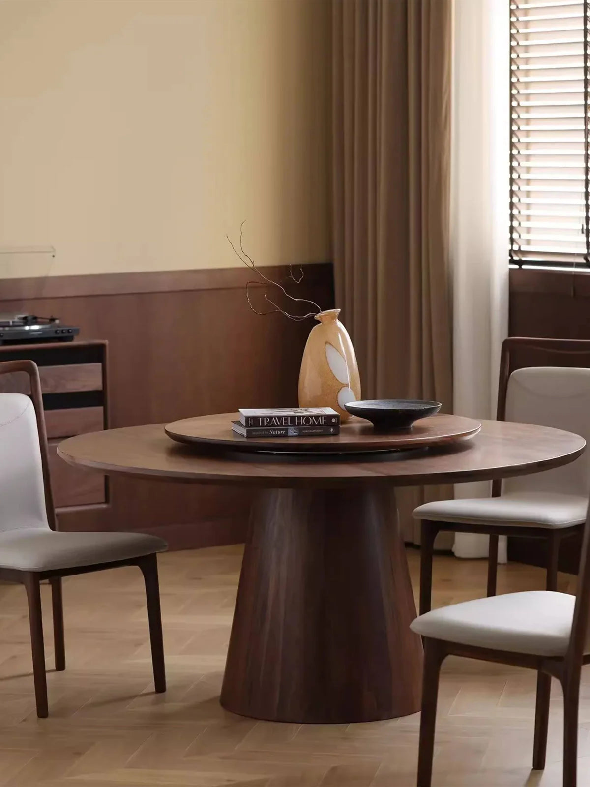 Round walnut pedestal dining table in a dining room setting with upholstered chairs, wood-paneled walls, and soft natural light.