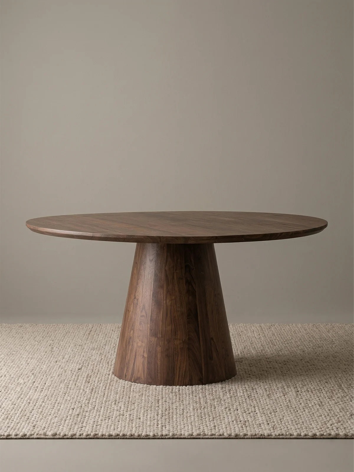 Studio view of a round walnut pedestal dining table with a thick tabletop and flared conical base on a textured neutral rug.