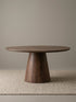 Studio view of a round walnut pedestal dining table with a thick tabletop and flared conical base on a textured neutral rug.