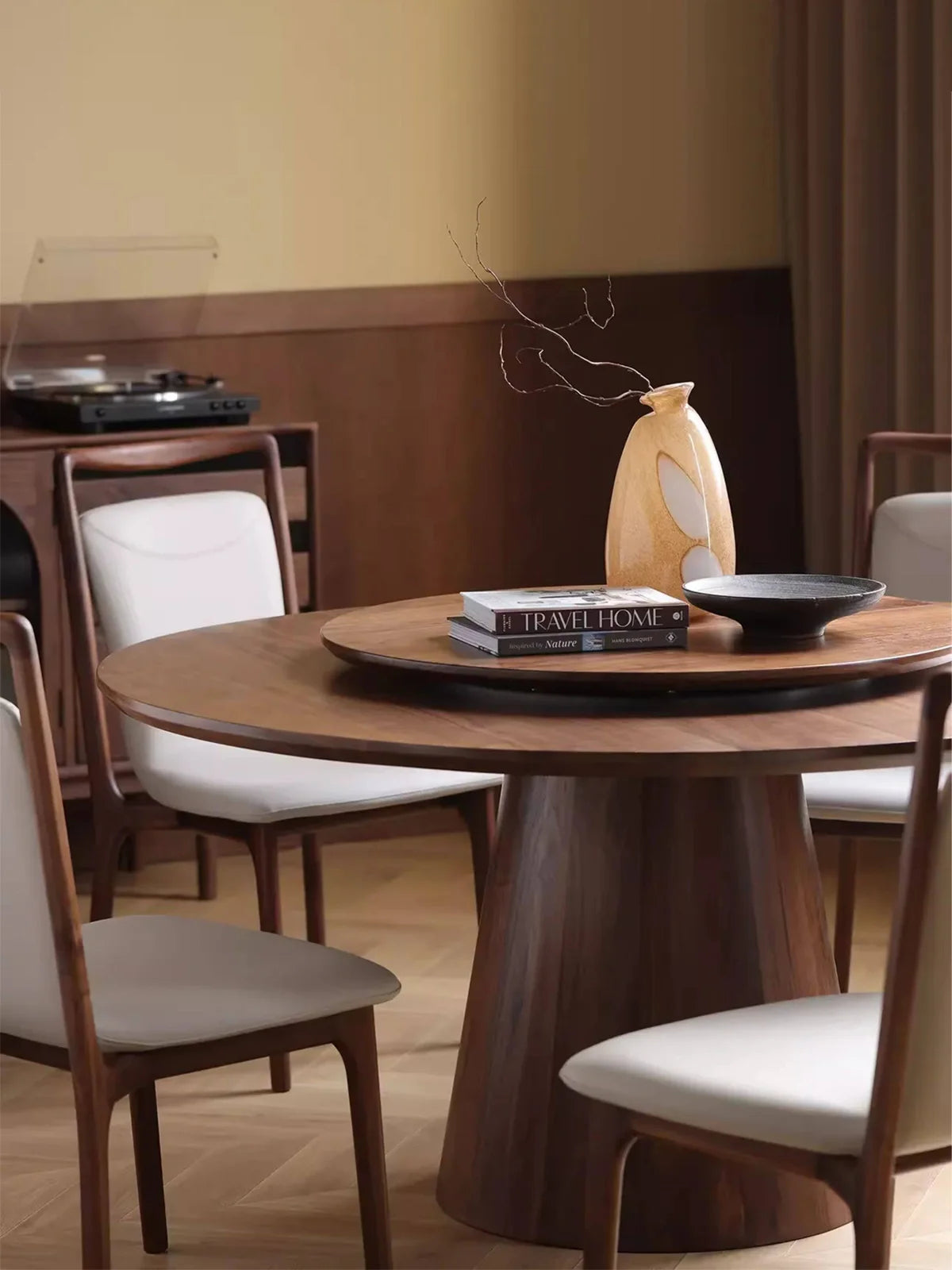 Three-quarter view of a dark walnut round dining table with a lazy Susan, styled with books, a vase, and upholstered dining chairs.