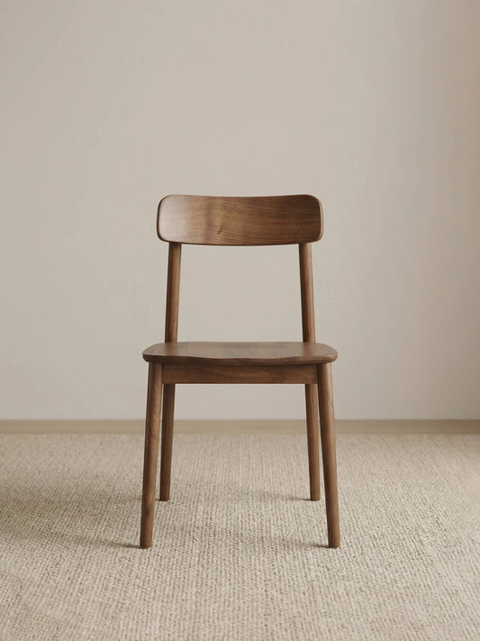 Front view of a solid American black walnut dining chair with a low backrest, rounded seat, and tapered legs on a neutral woven rug.