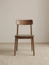 Front view of a solid American black walnut dining chair with a low backrest, rounded seat, and tapered legs on a neutral woven rug.