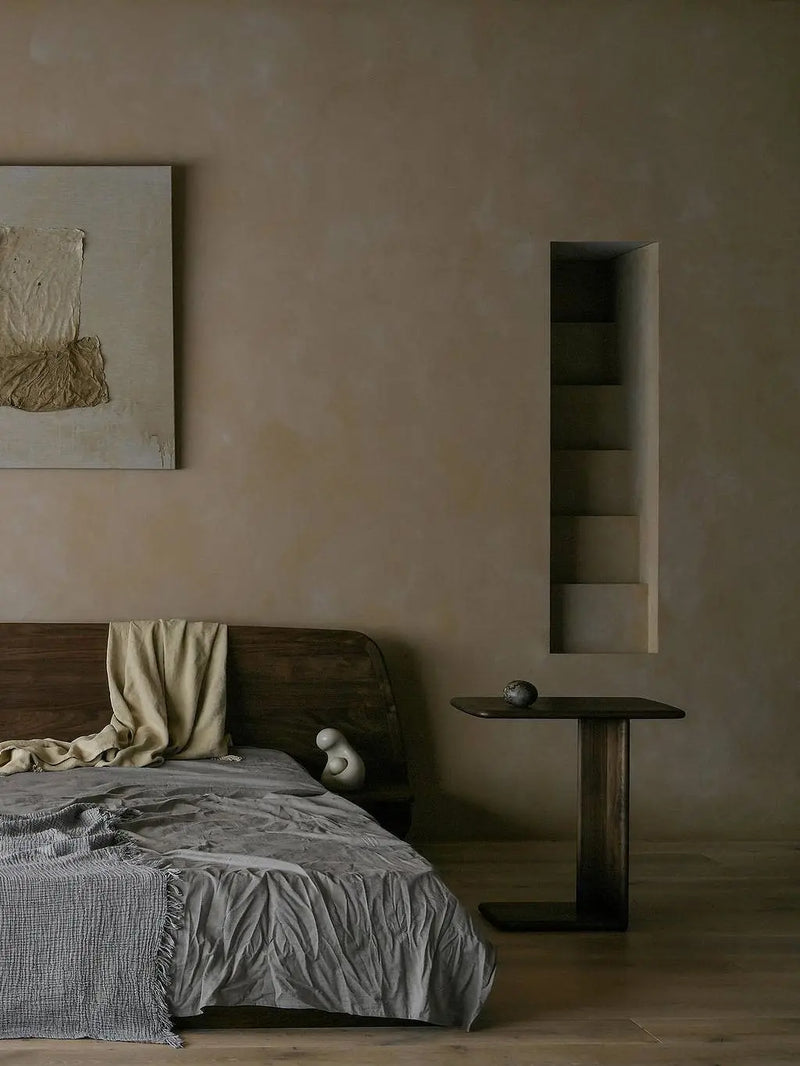 Walnutry solid North American Black Walnut bedroom furniture collection, featuring a minimalist low-profile platform bed and sculptural side table in a moody, wabi-sabi style bedroom with textured walls.