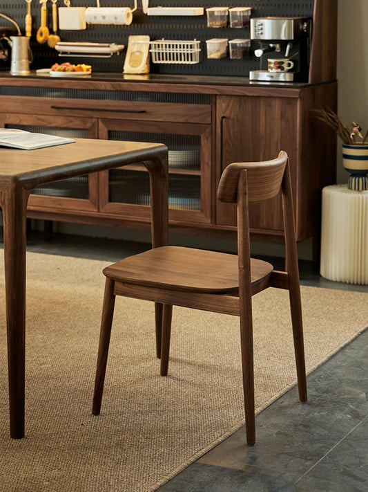 Solid walnut dining side chair styled beside a wood dining table in a warm kitchen interior, showing its slim profile and low back silhouette.