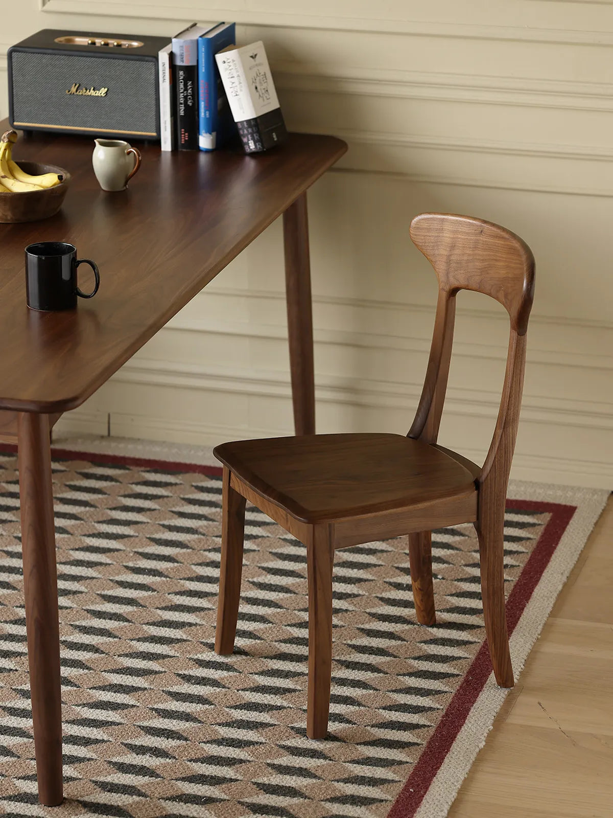 Sora dining chair in solid walnut tucked beside a walnut dining table in a styled room with books and a Marshall speaker