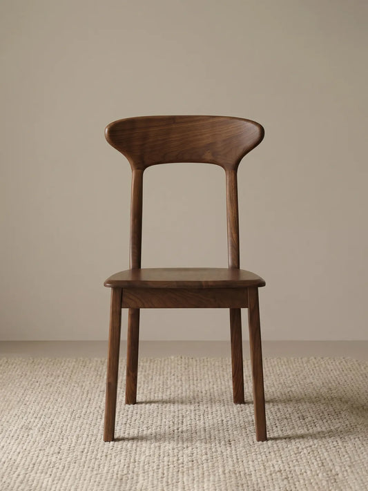 Sora solid walnut dining chair seen from the front, showing sculpted curved backrest and tapered legs on a cream rug