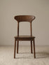 Sora solid walnut dining chair seen from the front, showing sculpted curved backrest and tapered legs on a cream rug