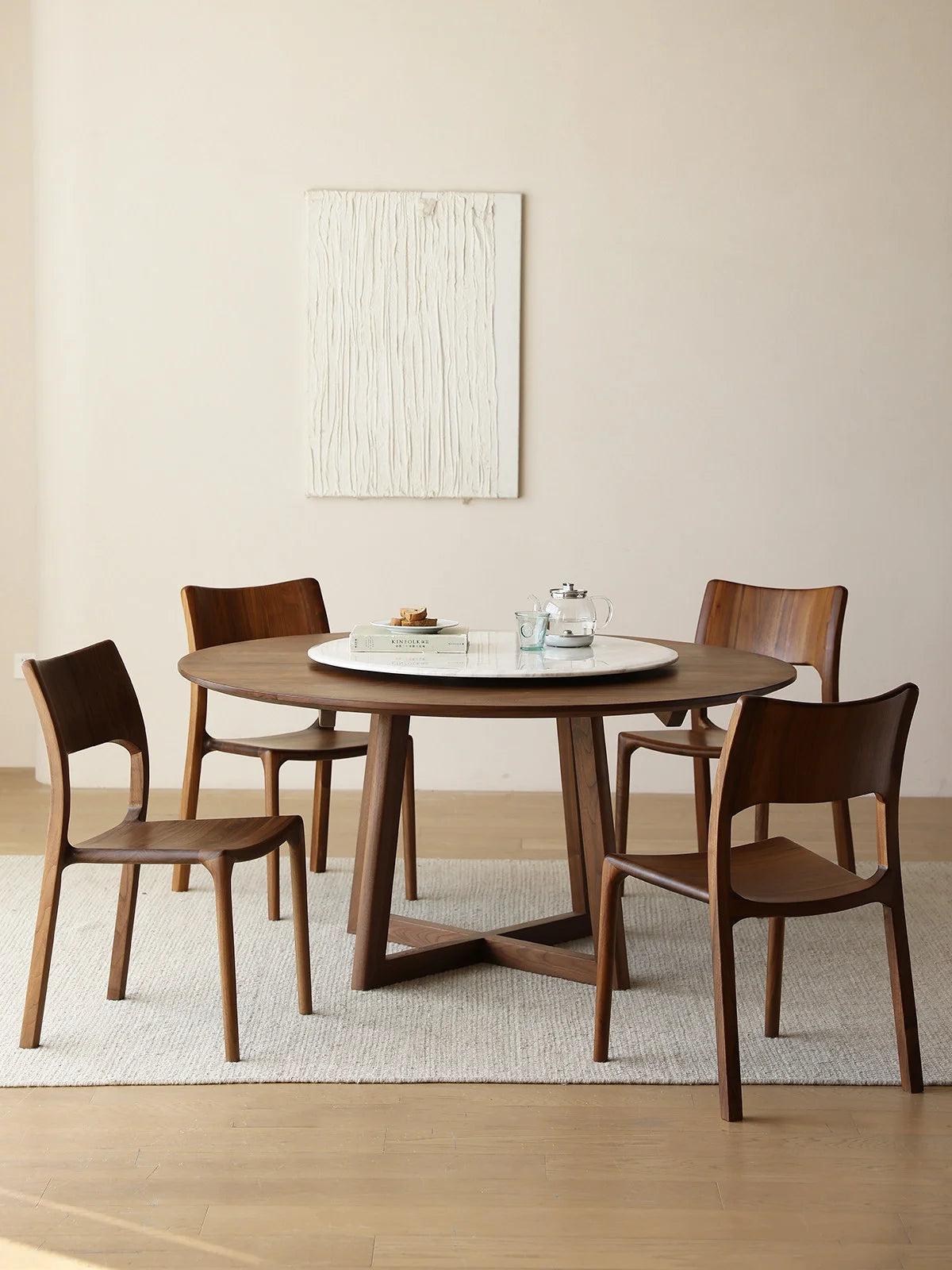 Front view of a round walnut dining table with four matching chairs and a white marble lazy Susan in a bright, pared-back interior.